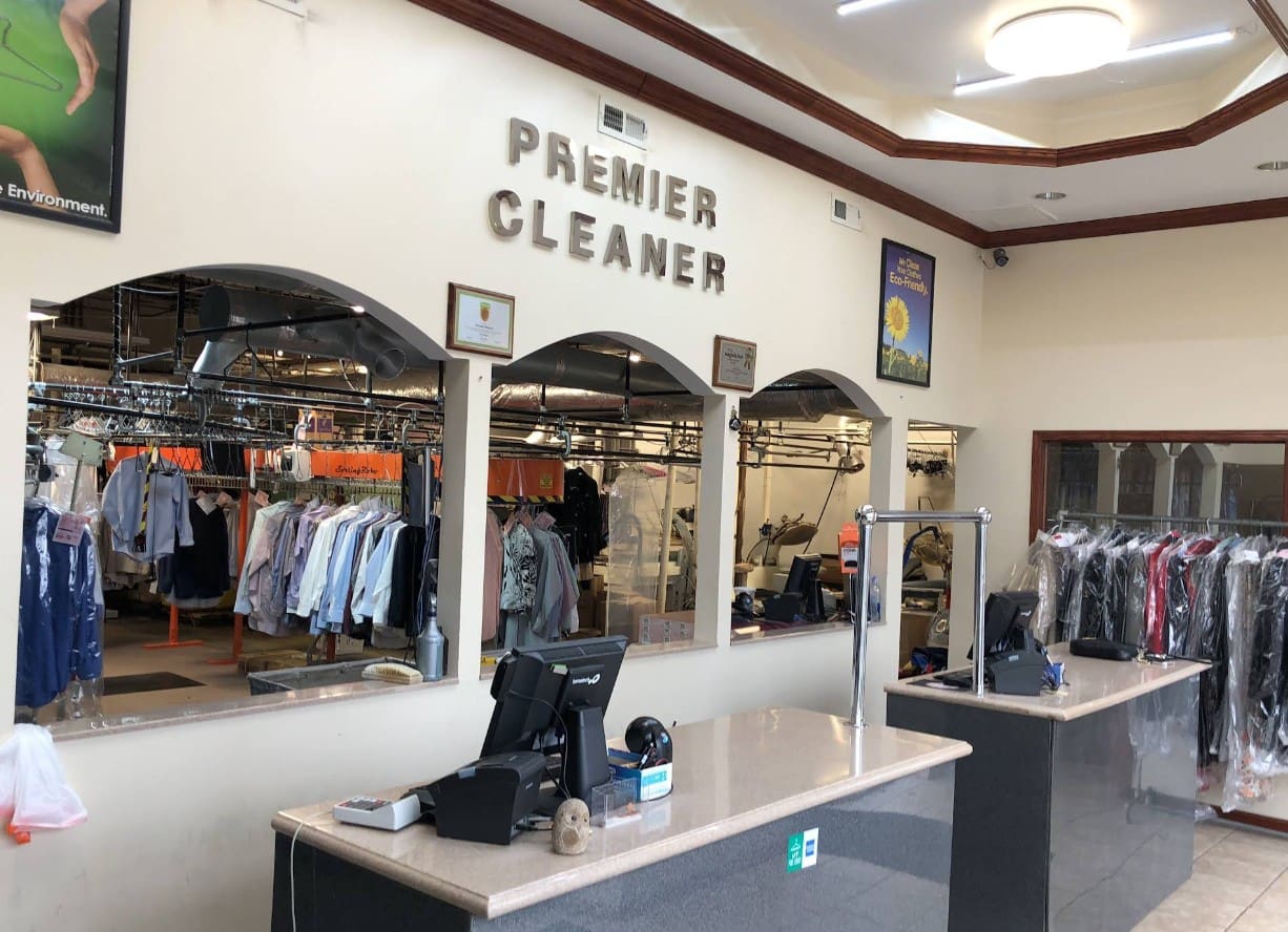 Interior of a dry cleaning shop with a front counter, cash register, mirrors, and racks of clothes visible behind the counter. The sign reads 