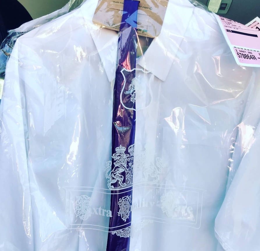 A white dress shirt on a hanger covered in plastic, with dry cleaning tags attached and a purple band with a white crest design across the front.