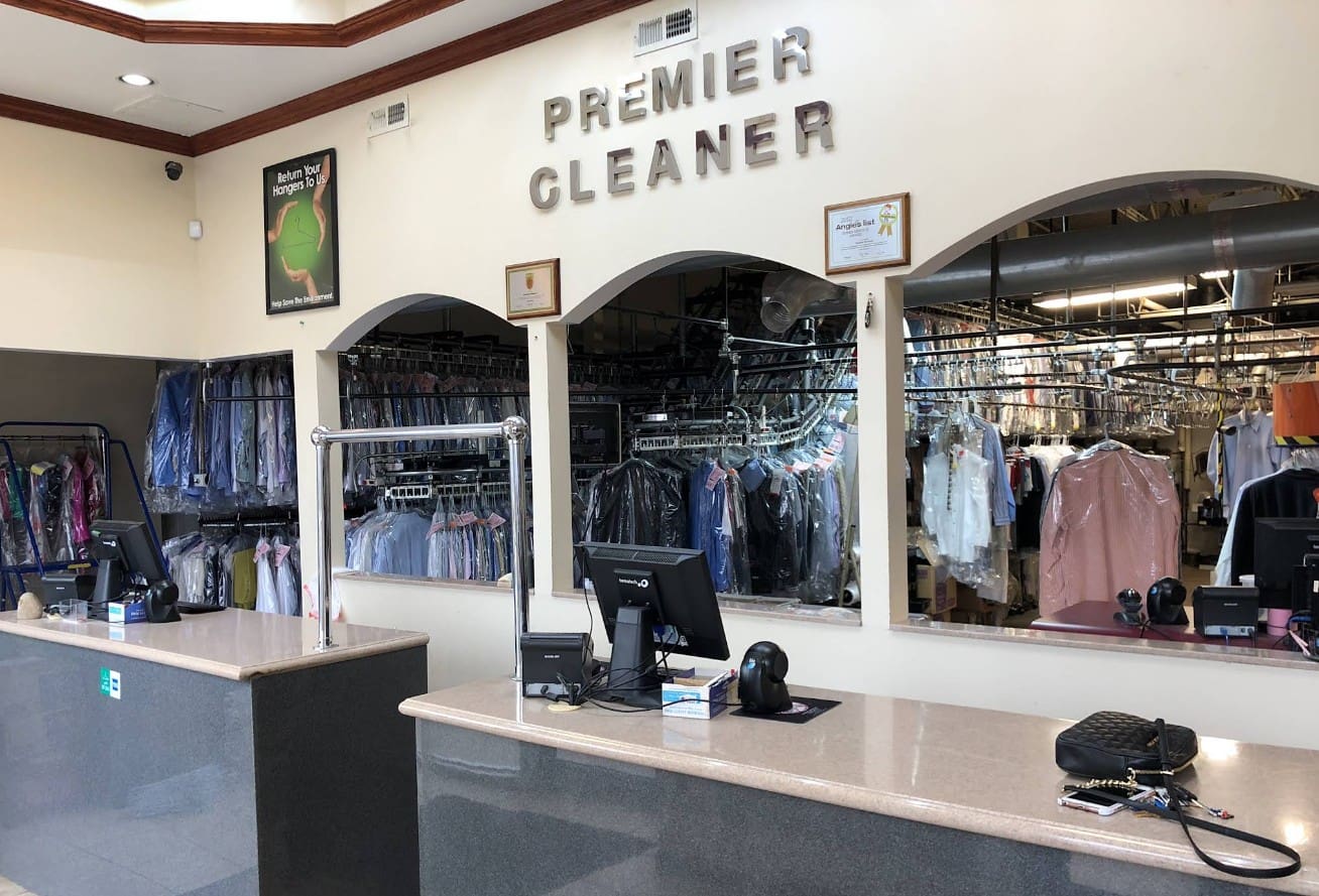 Interior of a dry cleaning business with two counters, computer terminals, and racks of clothes behind glass partitions. A sign above reads "Premier Cleaner.
