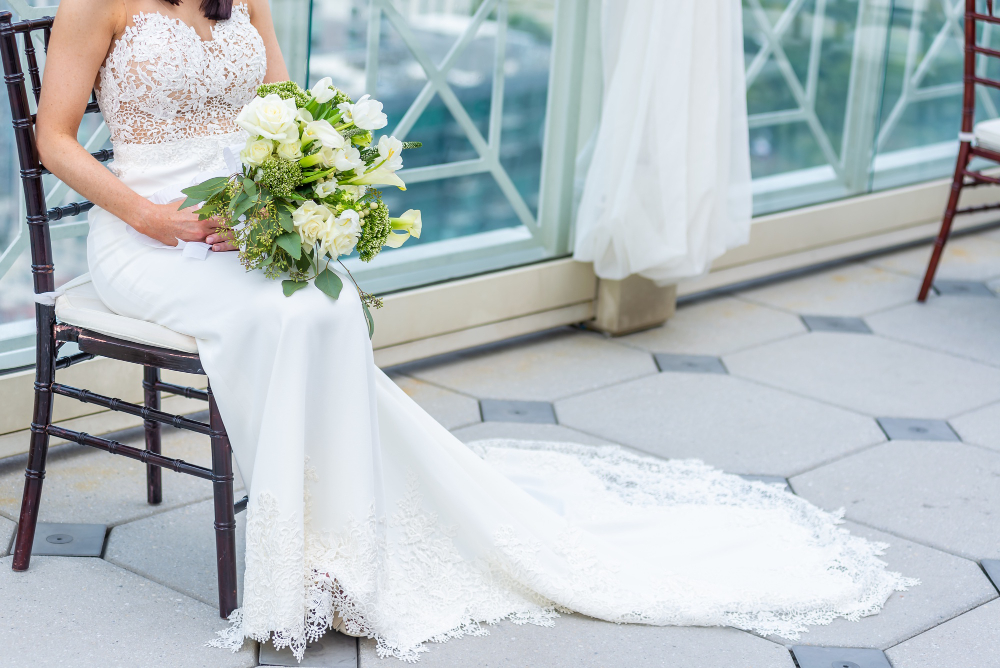 A bride in a white lace wedding dress sits on a chair, holding a bouquet of white and green flowers, with a long train spread on the floor.