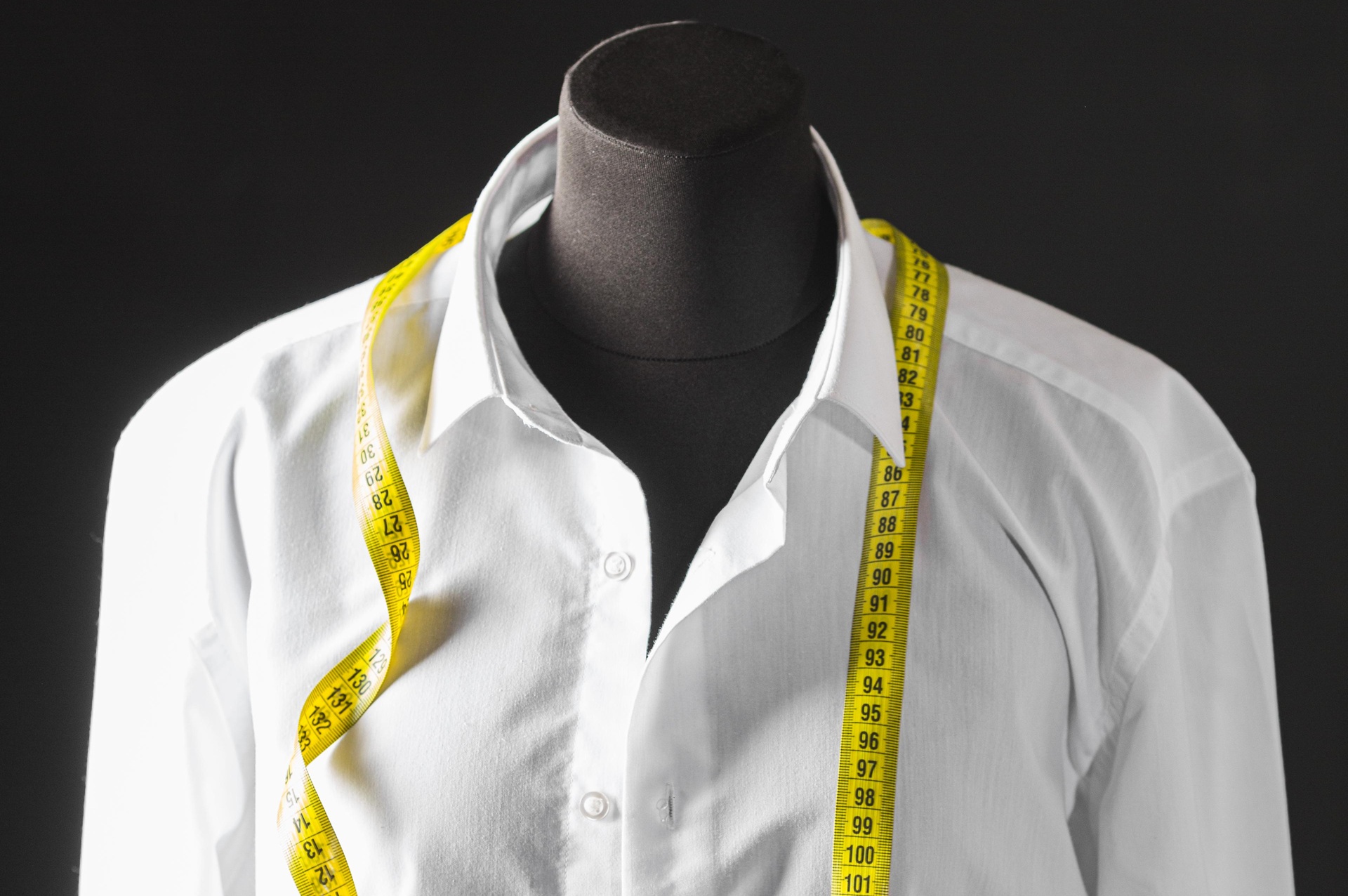 A black mannequin dressed in a white shirt with a yellow measuring tape draped around its neck against a black background.