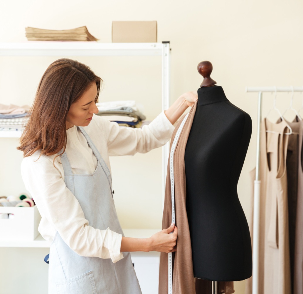 A woman in an apron drapes fabric on a black dress form and measures it with a tape measure in a workspace with shelves and hanging garments.