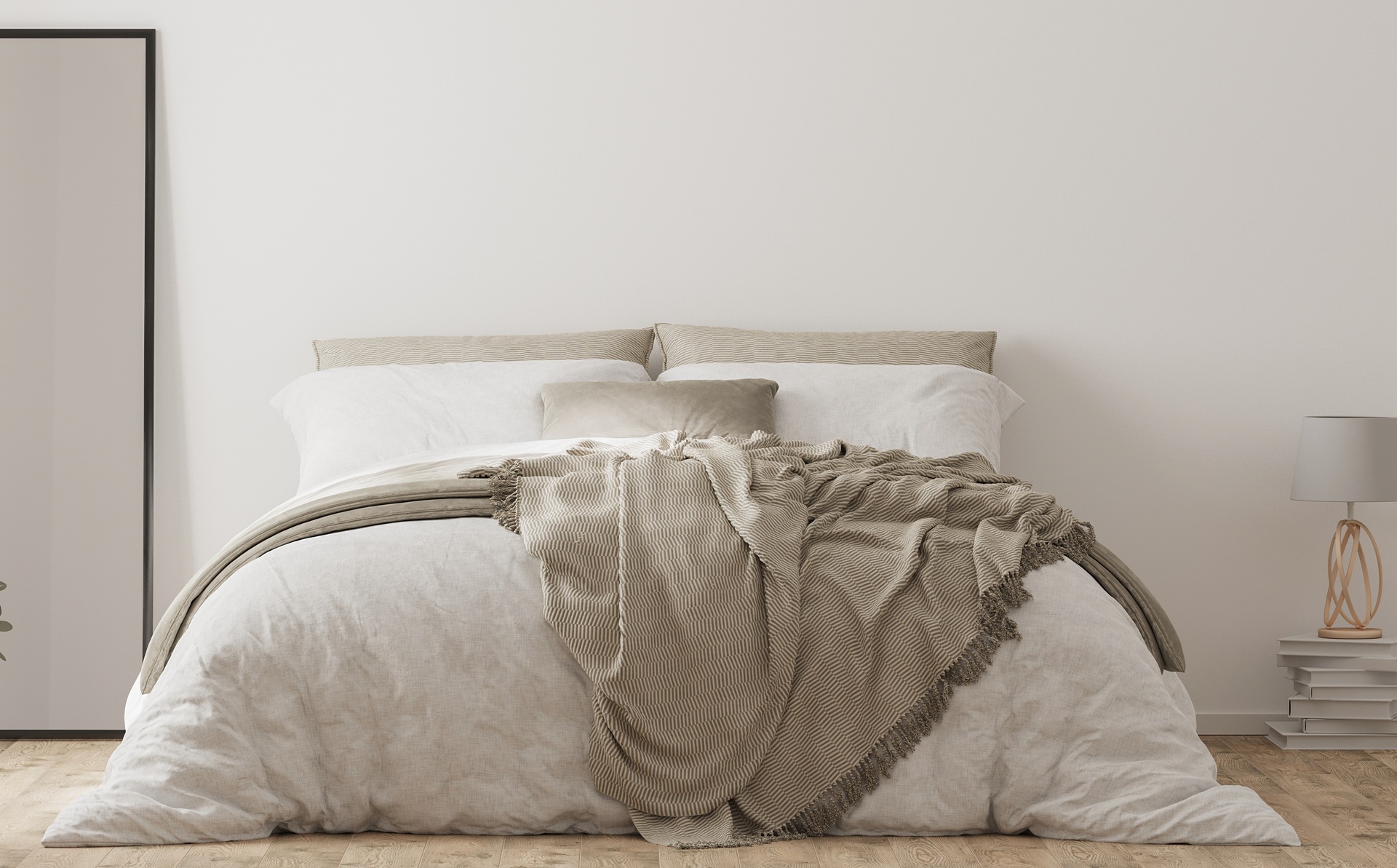 A neatly made bed with white bedding and beige cushions, a textured beige throw blanket, a floor mirror, a small lamp, and stacked books on a nightstand in a minimalist bedroom.