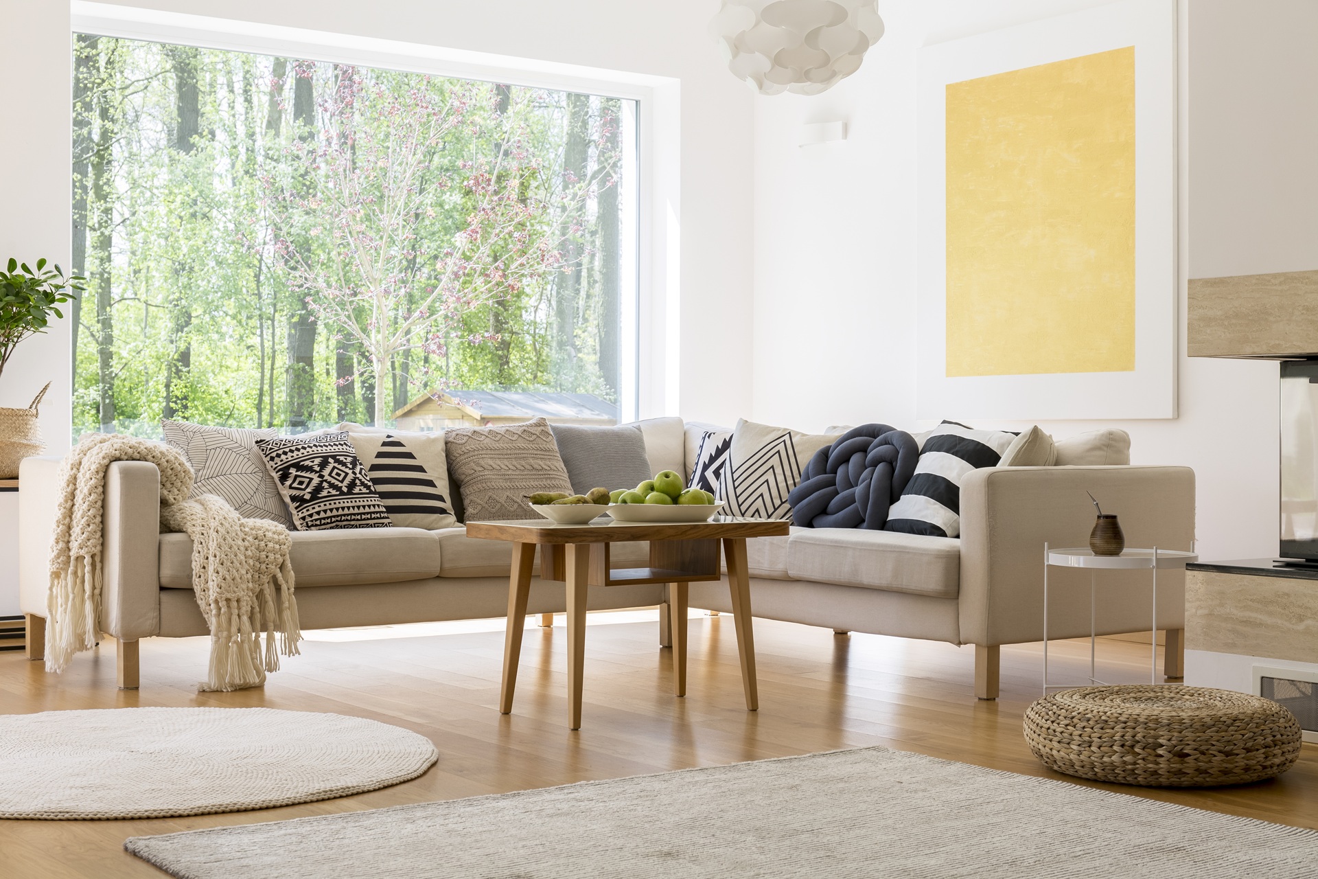 A modern living room with a beige sofa, patterned cushions, a wooden coffee table with apples, large window with a forest view, and minimalist decor.