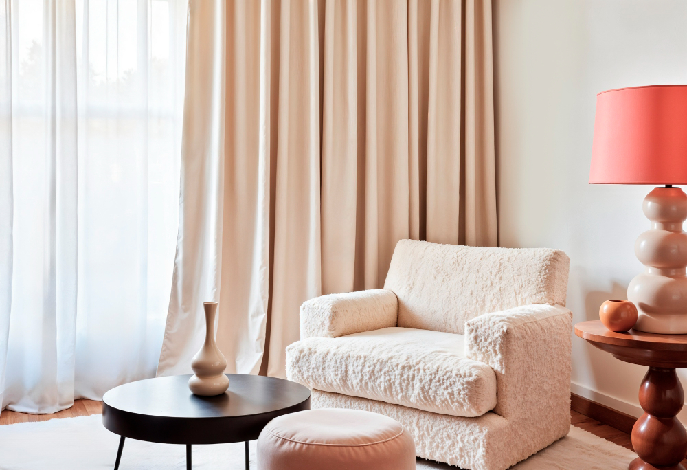 A cozy living room with a textured cream armchair, round black coffee table, beige ottoman, pink lamp on a wooden side table, and beige curtains covering a window.