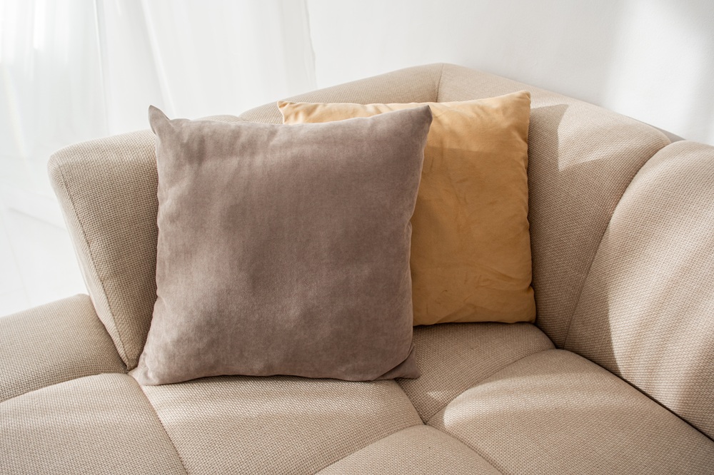 Two square pillows, one beige and one light brown, are placed on the corner of a light-colored fabric sofa in a bright room.
