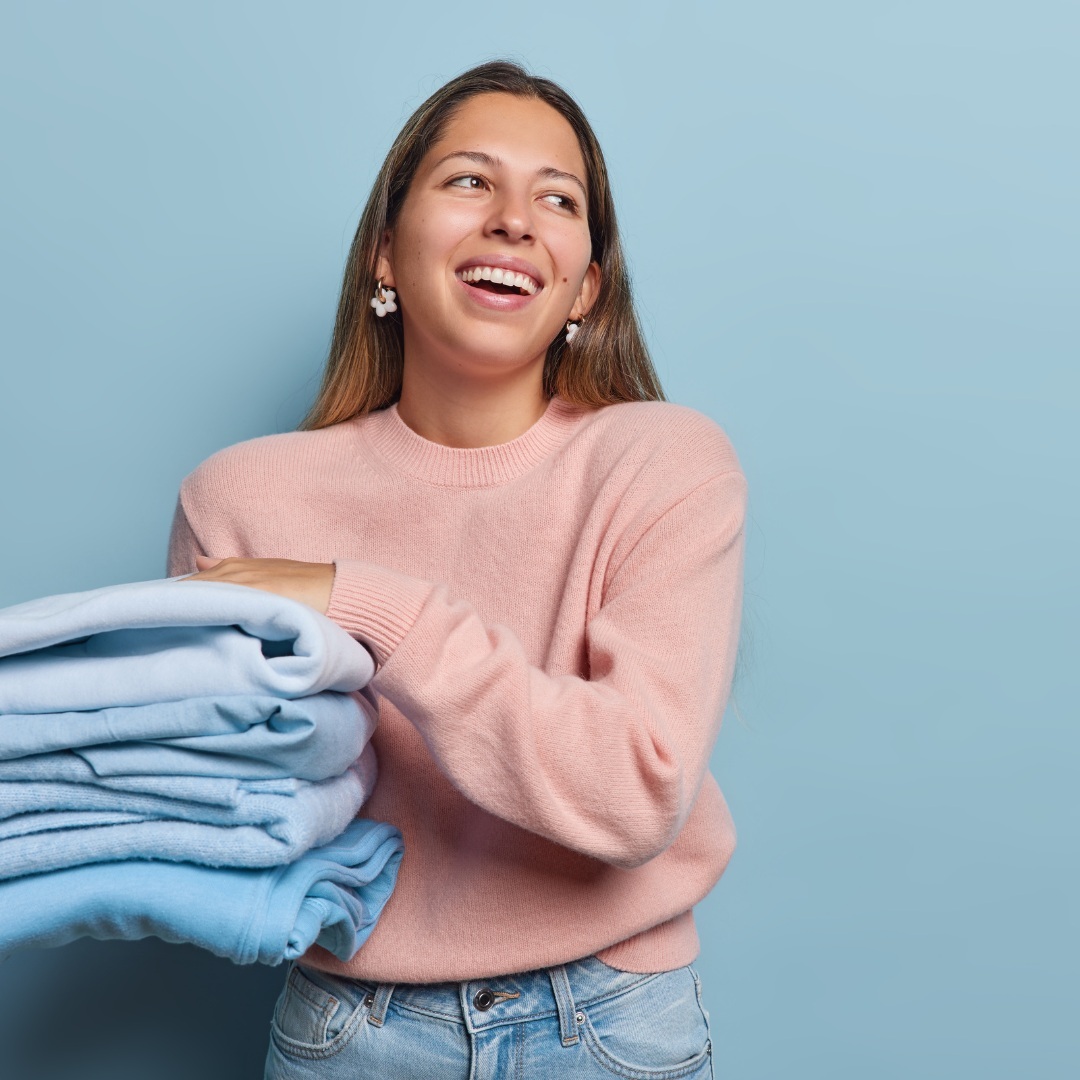 A woman wearing a pink sweater holds a stack of folded blue clothes and smiles, standing against a plain light blue background.