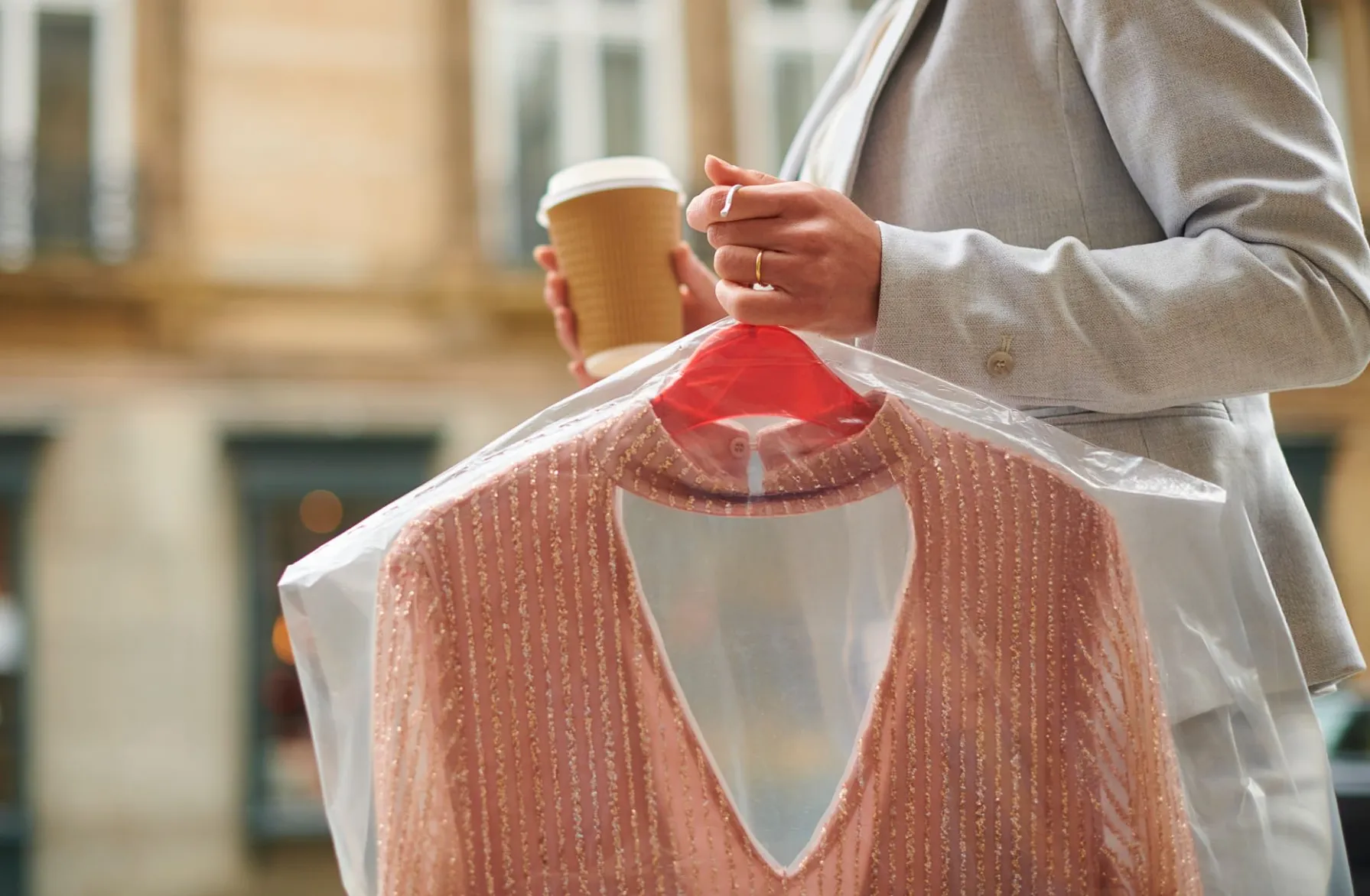 Person holding a coffee cup and a hanger with a pink beaded dress covered in a plastic garment bag, standing outdoors.