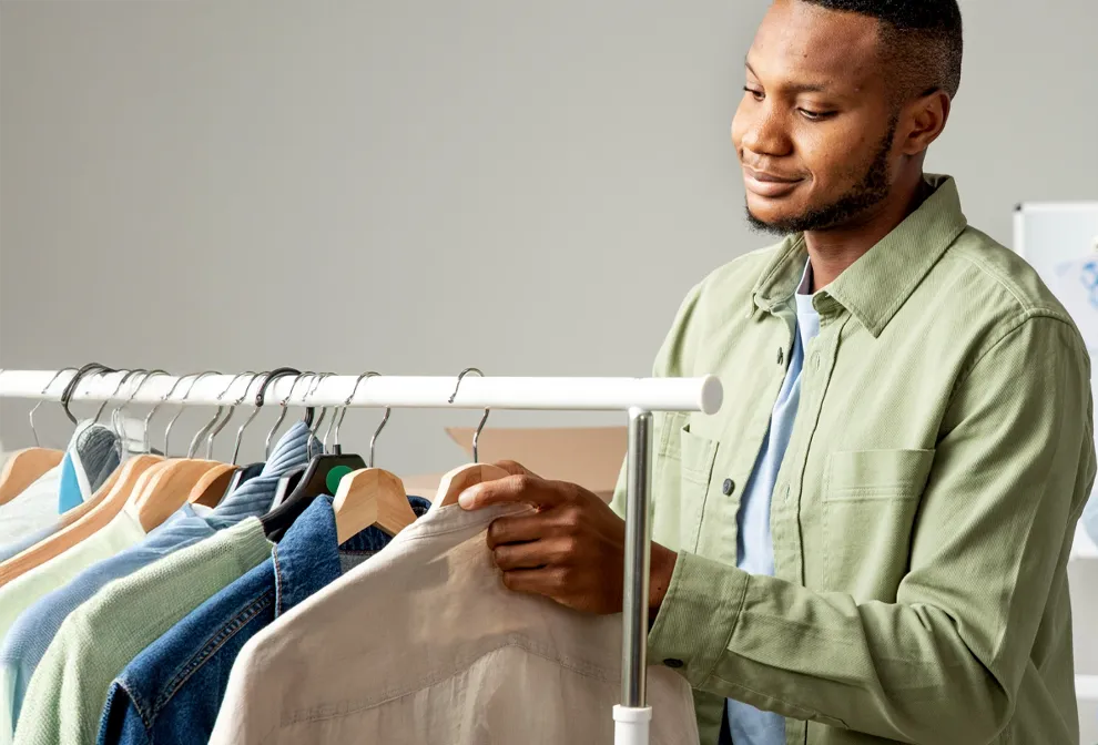 A man in a green shirt looks at and arranges clothing on a rack, with several shirts hanging on wooden and metal hangers.